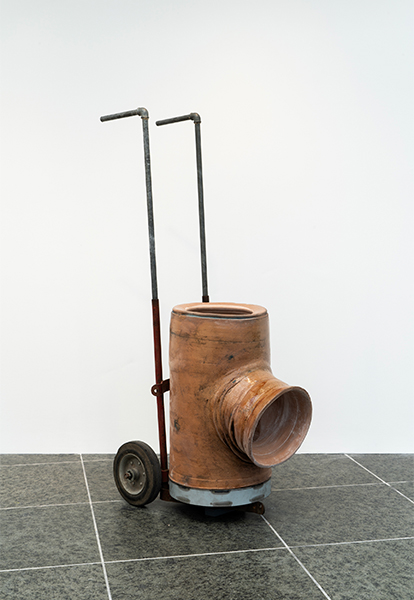 A minimal hand truck supports a large, handmade, terracotta ceramic sewer pipe. It  resembles a T-shaped pipe fitting, with three openings.