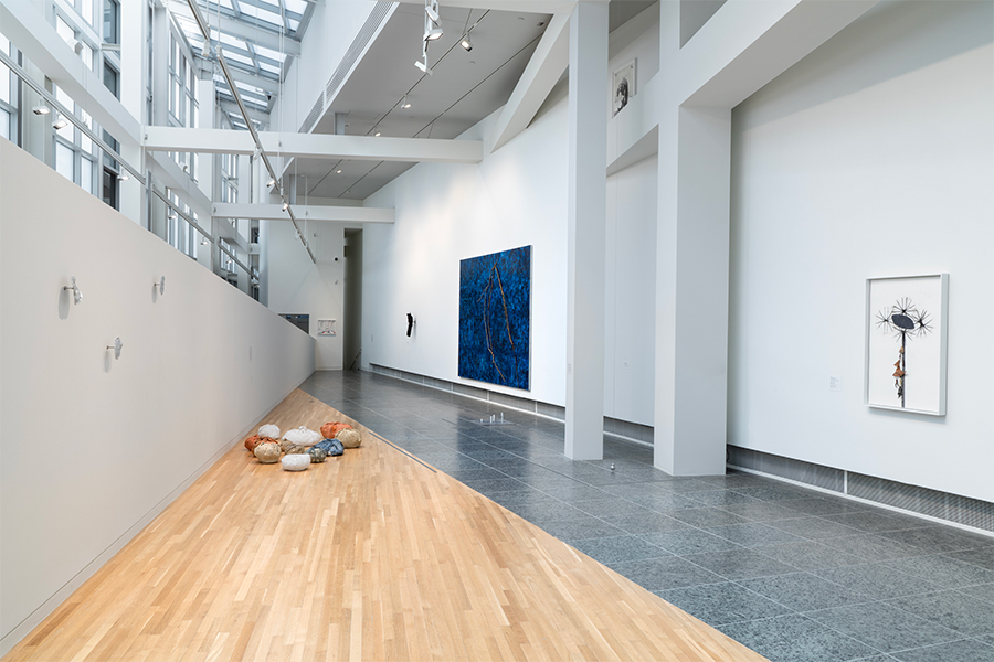 Mixed-media wall works hang to the left of multi-colored bundle shaped ceramic sculptures on the floor. The triangular gallery architecture, windows, and beams are visible.