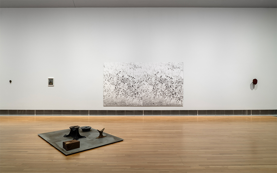 Four mixed-media wall works are arranged left to right on a white wall. Five-piece mixed-media sculpture rests on a metal plate on a wooden floor.