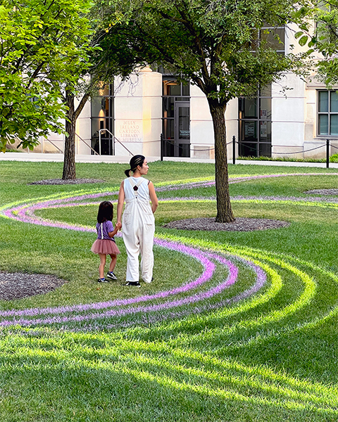 An adult and child walk on a lawn painted with pink, yellow, and white lines. The lines curve around the trees and overlap one another.