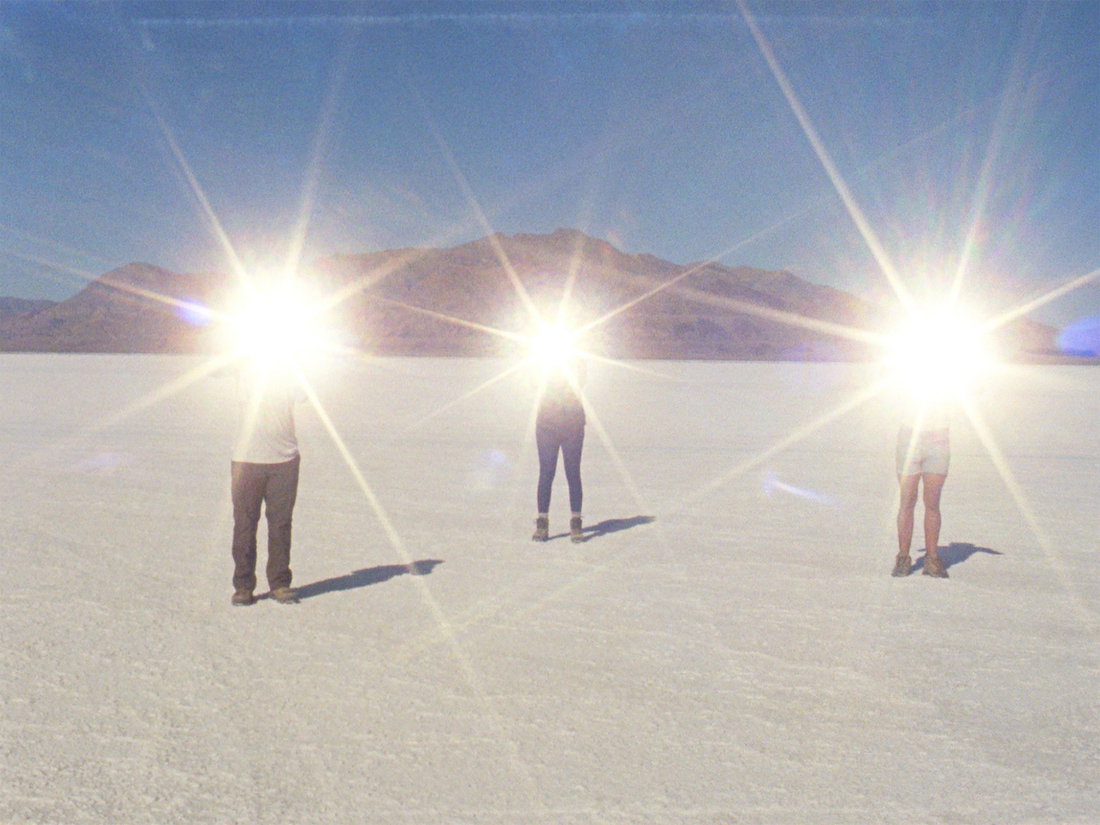 Three people stand apart in a salt flat environment while holding mirrors towards the sun, reflecting brightly and obscuring their faces.