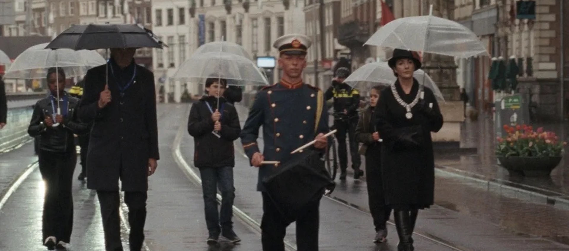 A group of six people of various ages walk down a rainy city street, most with umbrellas over their heads.