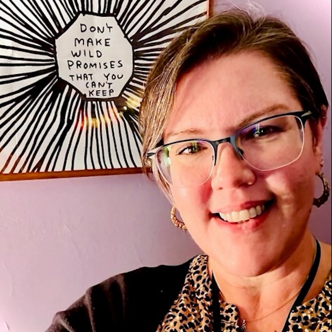 A woman with short brown hair and glasses smiles warmly as she stands in front of a wall hanging that reads, “Don’t make wild promises that you can’t keep.”