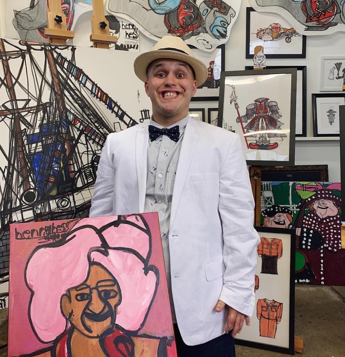 A young man smiles as he stands in a full artist studio space with a portrait painting in his hand.