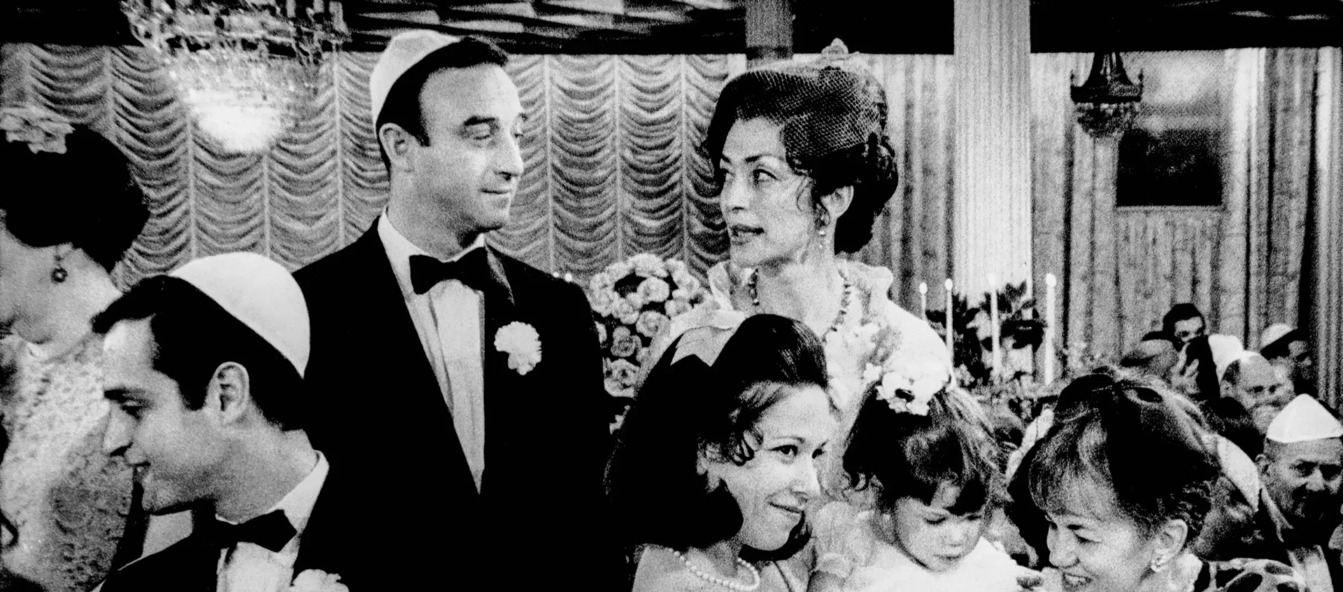 A large group of people gathered at a fancy party. The men are all wearing yarmulkes and tuxedos and the women are wearing dresses. 