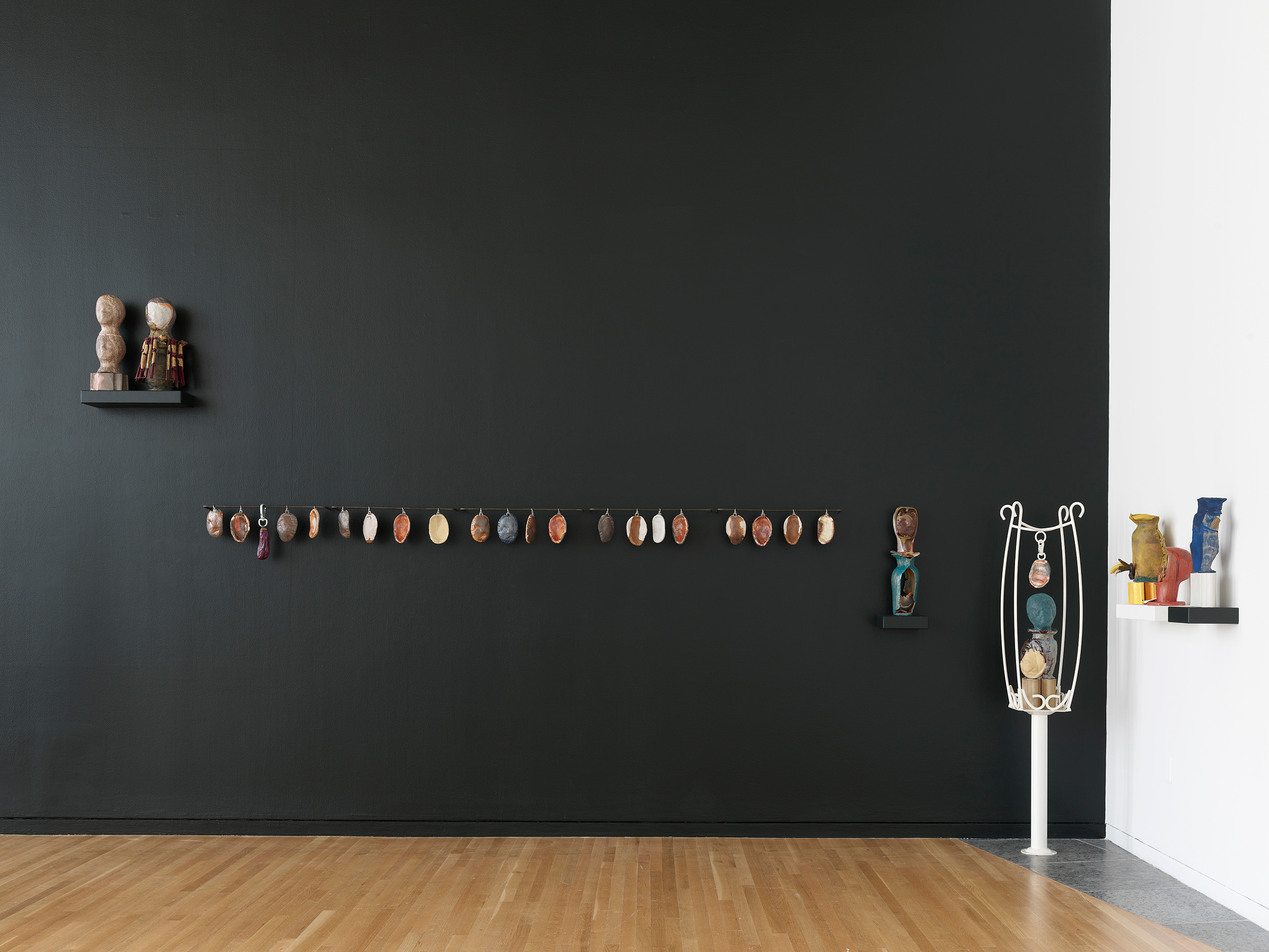 Figurative sculptures on a shelf above a row of earth-toned ceramic pitas, next to multicolored vases and a white columnar and wire pedestal holding sculptures.
