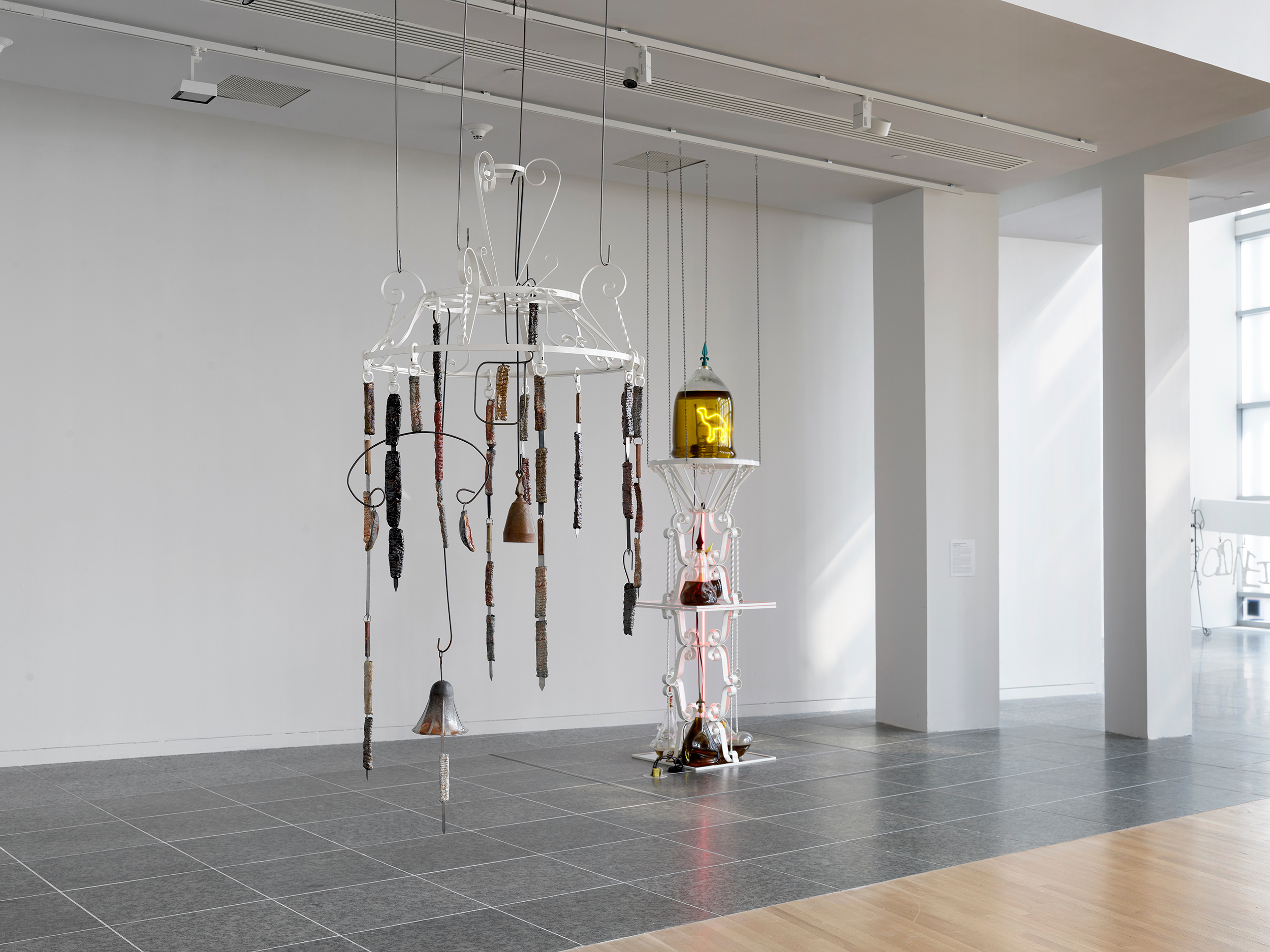 Installation of two sculptures made of metal, ceramics, and glass suspended from the ceiling and resting on a divided wood and granite floor. 