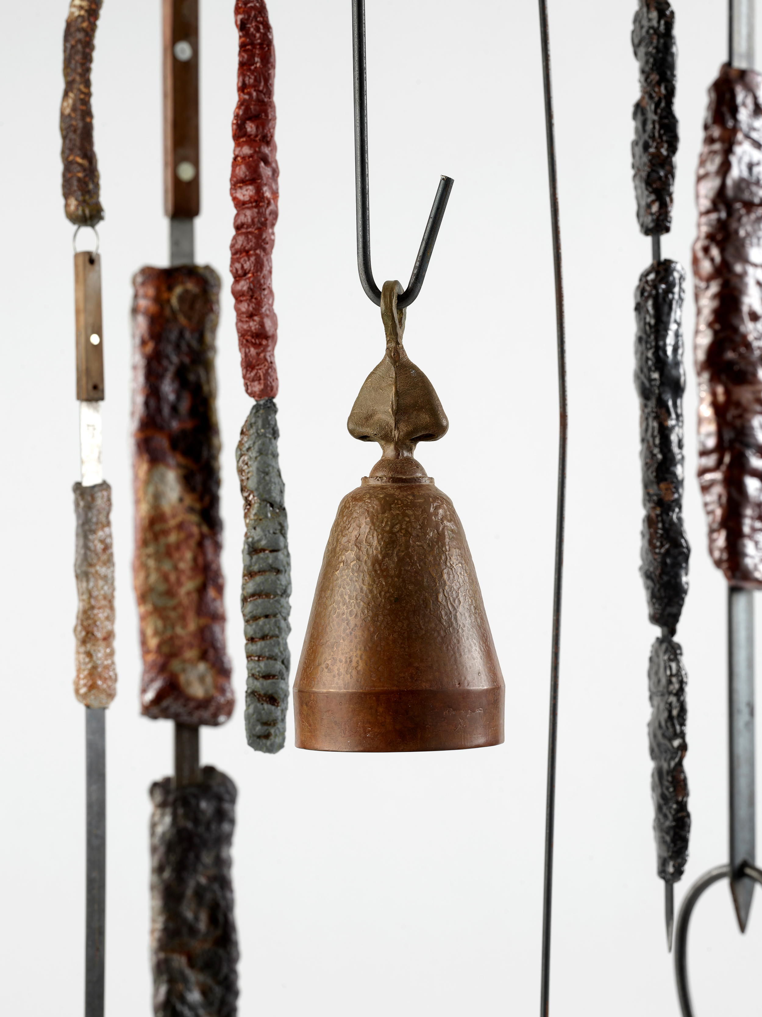 Detail of earth-toned sculpted ceramic kebabs and a conical bell topped by a sculpted nose hanging from a wind chime frame. 