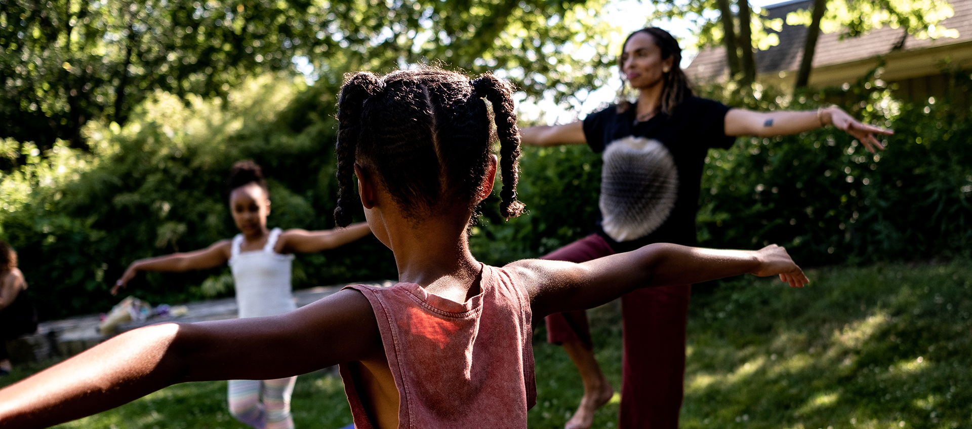 Two children and an adult with outstretched arms balance on one foot on yoga mats in an outdoor setting.