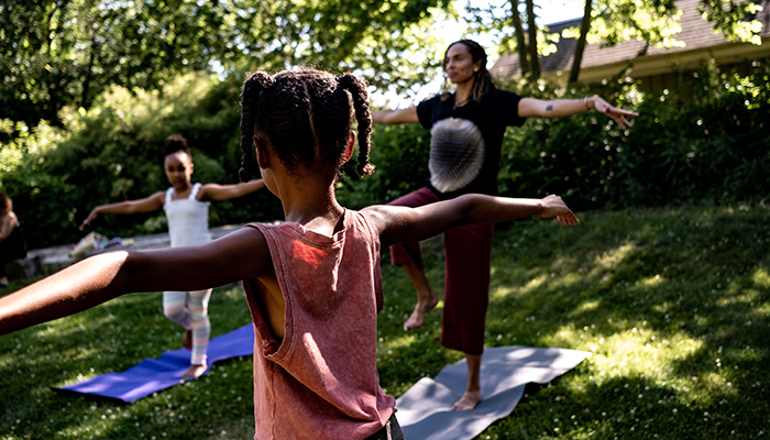 Two children and an adult with outstretched arms balance on one foot on yoga mats in an outdoor setting.