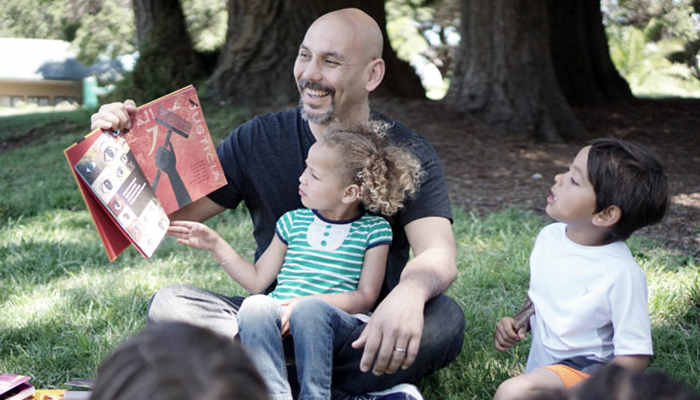 Smiling bald man with mustache and beard reading a book to children outdoors under the trees.