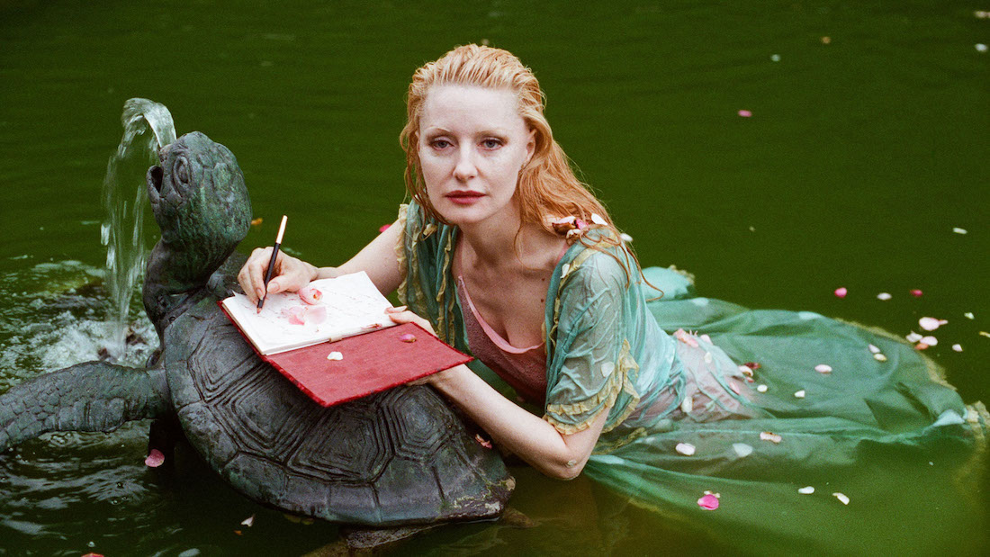 A woman looks at the camera as she lies in a pond. She leans forward on a turtle-shaped fountain and writes in a diary.