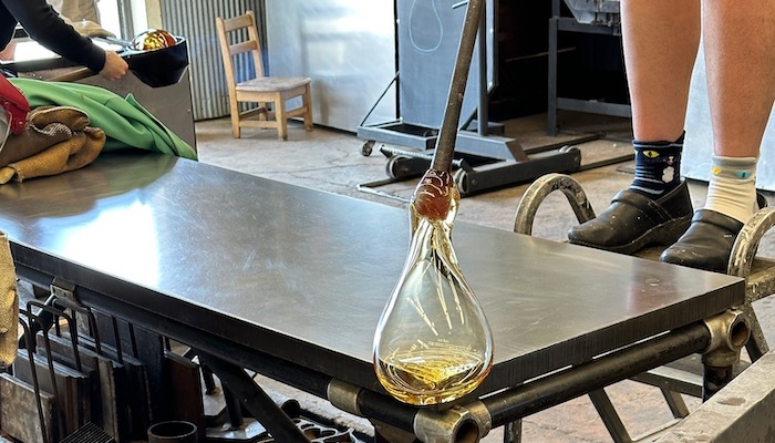 A teardrop-shaped clear glass form hangs from a metal pole over the edge of a metal table. The person holding the pole is partially visible.
