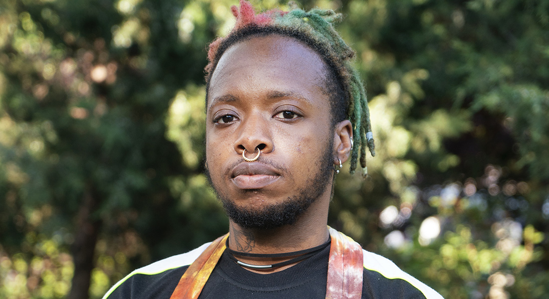 A brown-skinned person with multicolored, pulled-back dreadlocks faces the camera against a tree-filled background.