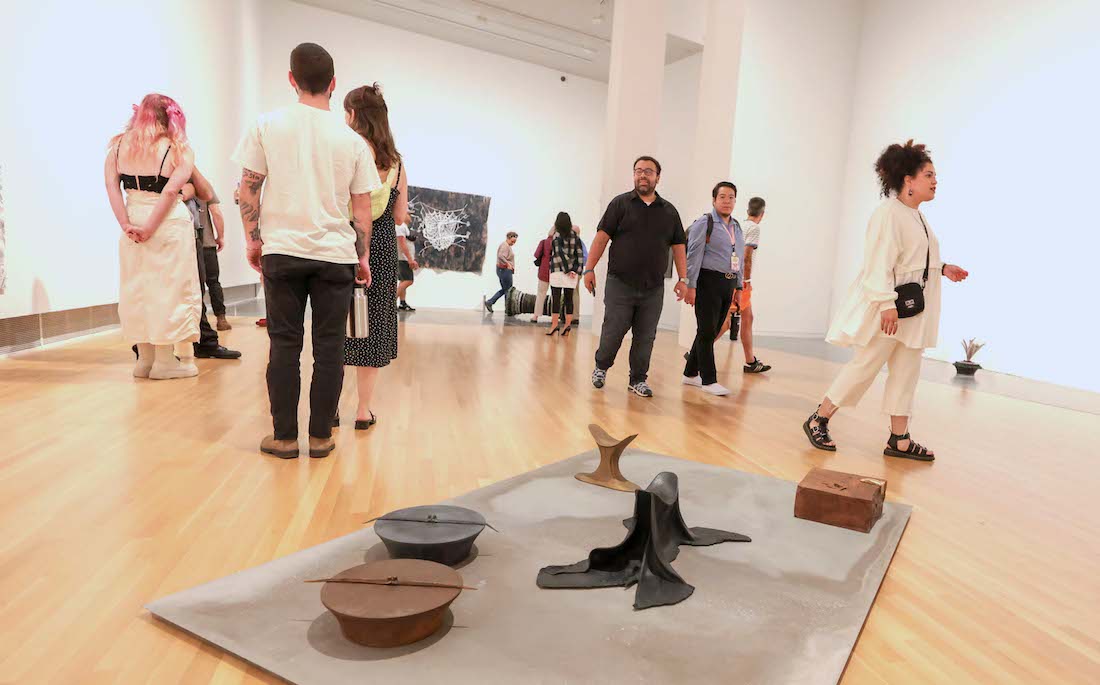 About 10 people are scattered and look around a gallery. A sculpture installation sits on the floor in the foreground.