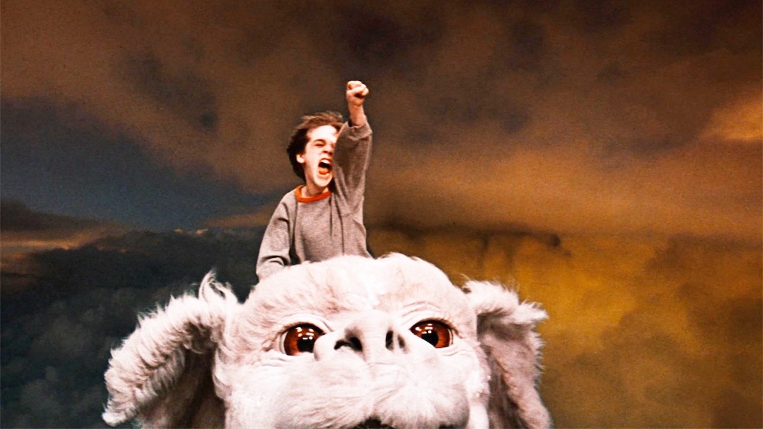 A young boy yells and raises his fist in triumph as he rides a large flying dog.