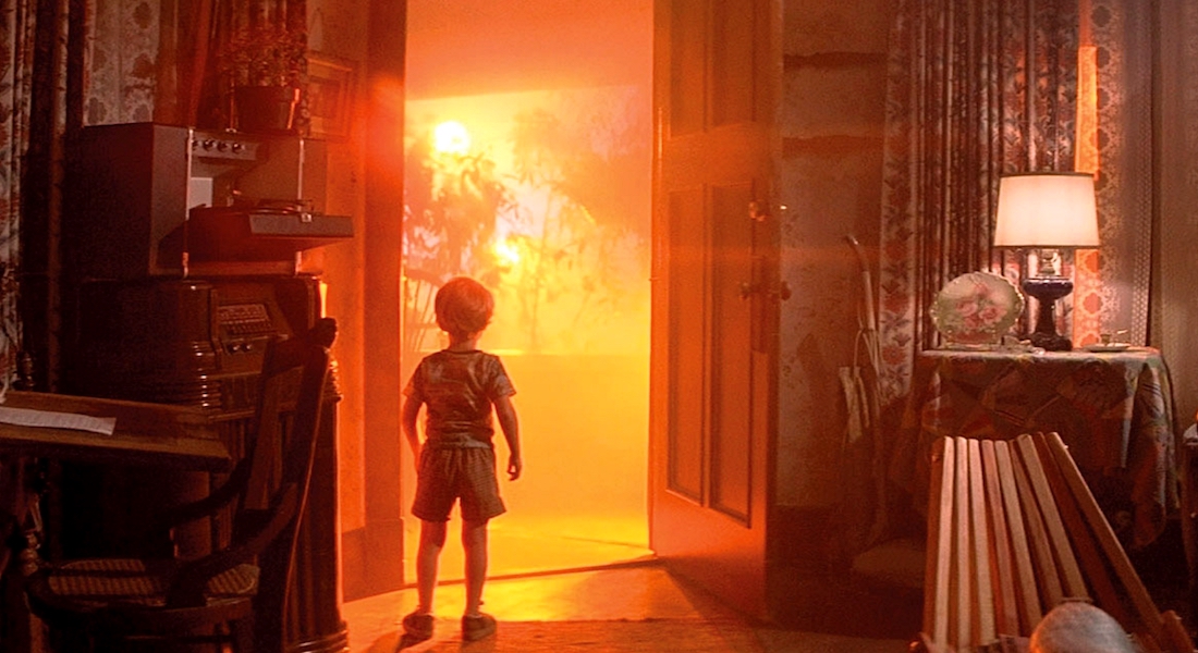 A little boy is standing in an open doorway, from which an orange light emanates.