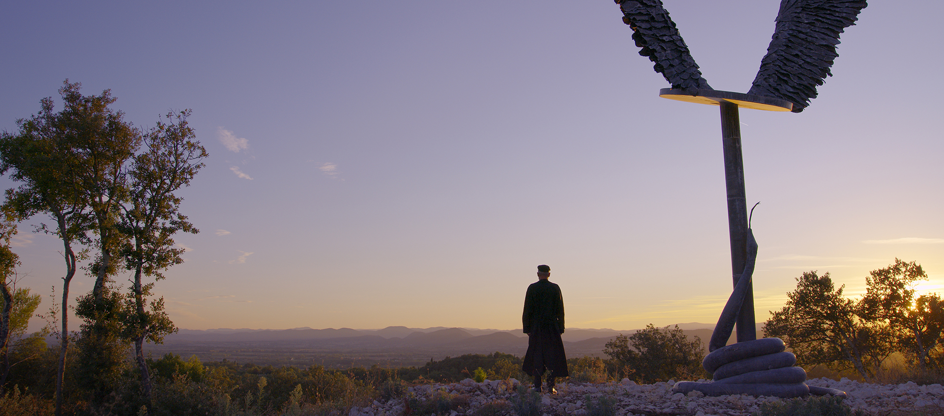 A person stands facing mountains at sunset, next to a large statue of angel wings on a platform with a snake spiraling up the pole underneath it.