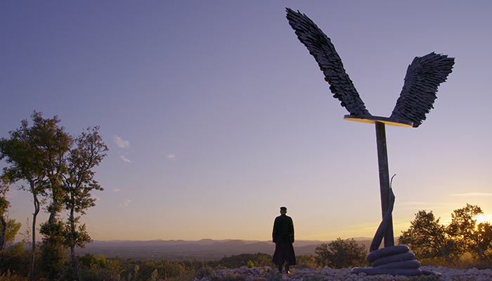 A person stands facing mountains at sunset, next to a large statue of angel wings on a platform with a snake spiraling up the pole underneath it. 