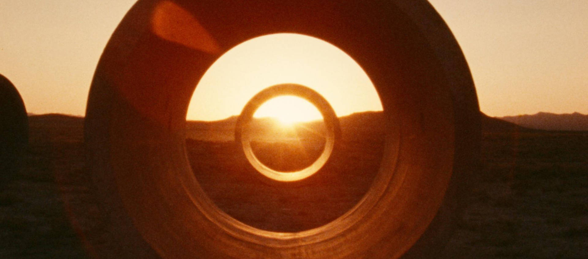 The orange sun setting over the desert landscape framed in a circle by two concrete cylinders.