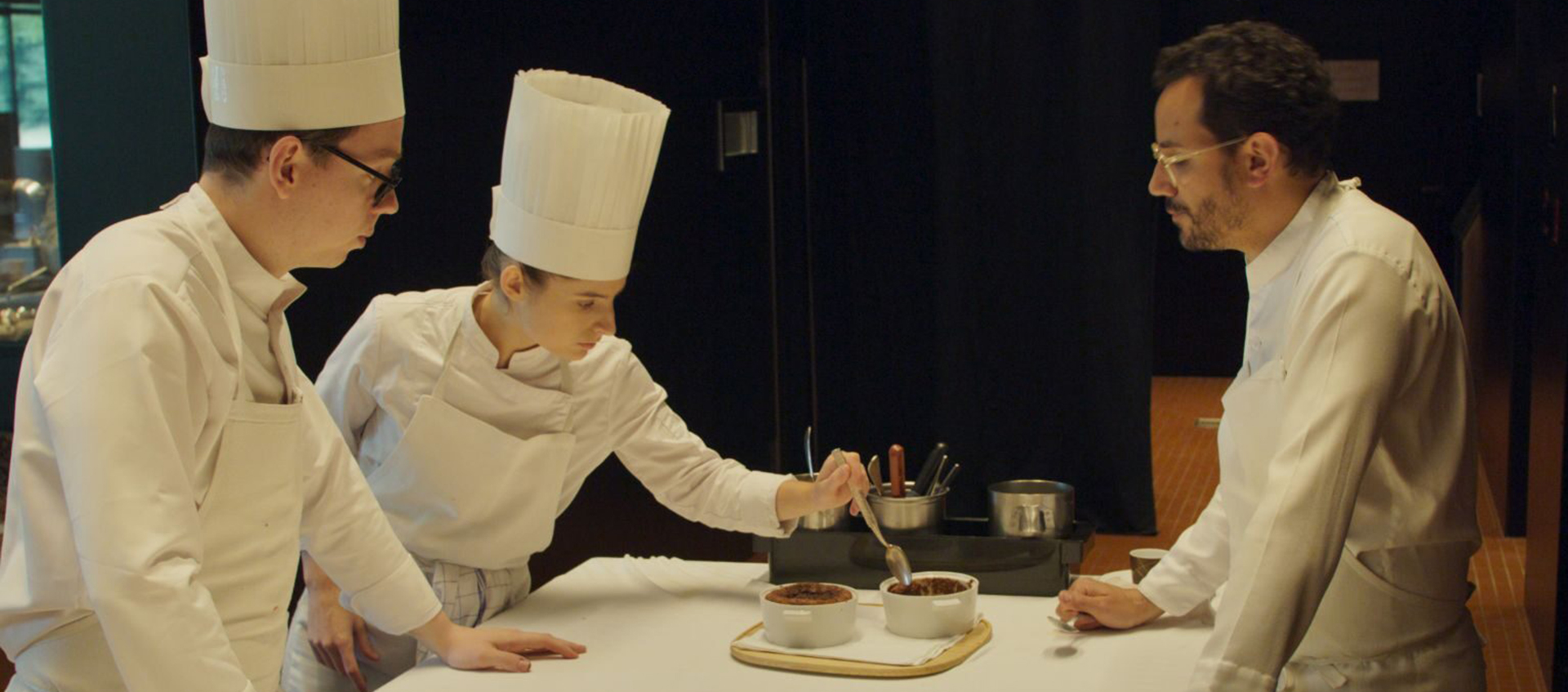 Three individuals are standing around a counter, two are wearing chef's hats.