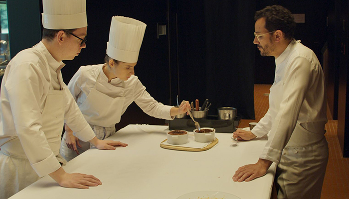 Three individuals are standing around a counter, two are wearing chef's hats.