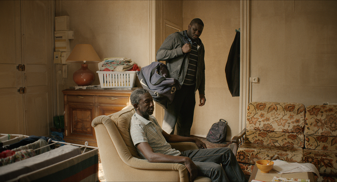 An older Black man sits in a living room chair. A younger Black man holding a duffel bag stands behind him, looking down at him.