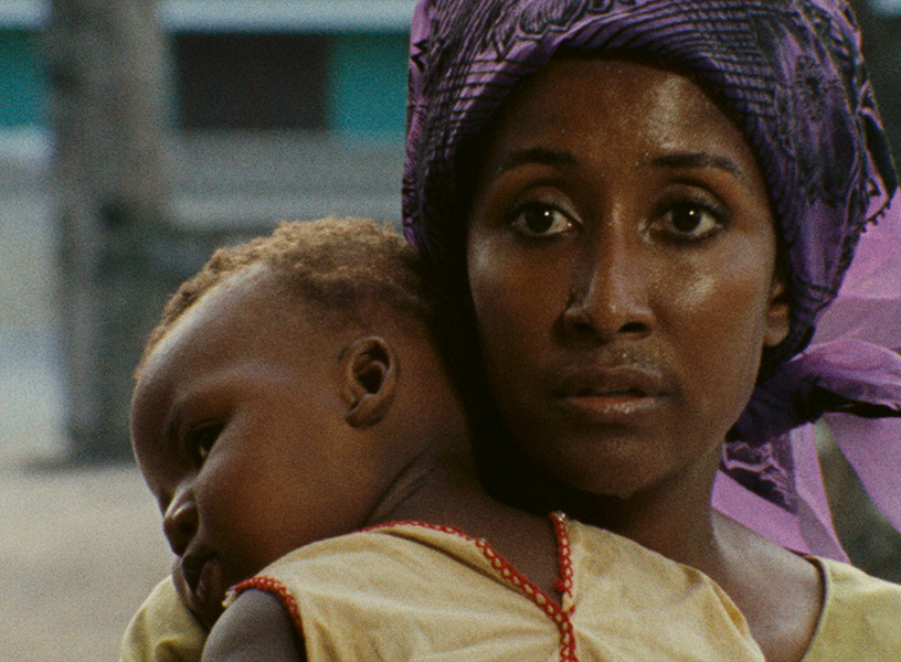 Close up of a young Black woman’s face, looking into the camera. She wears a blue head covering and holds a baby over her shoulder.