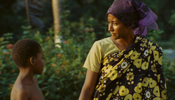 A young Black woman wearing a blue head covering and floral wrap dress talks to a shirtless young Black boy standing to her right. 