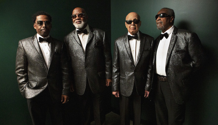 Four adults with brown skin wearing silvery suits, bow ties, and sunglasses face the camera in a row before a dark green wall.  