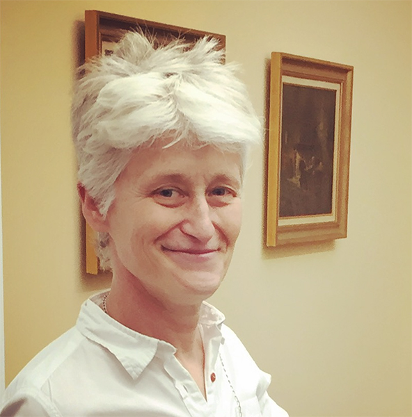 Elizabeth Povinelli smiles at the camera. She has light skin, short white hair, and wears a white shirt. Two paintings hang behind her.