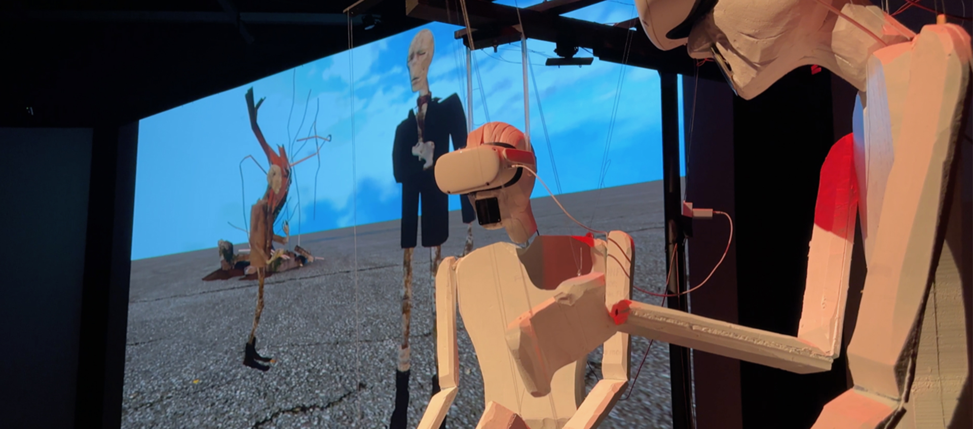 Two marionette robots wearing virtual reality visors stand in front of a screen