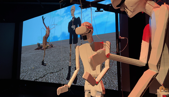 Two marionette robots wearing virtual reality visors stand in front of a screen