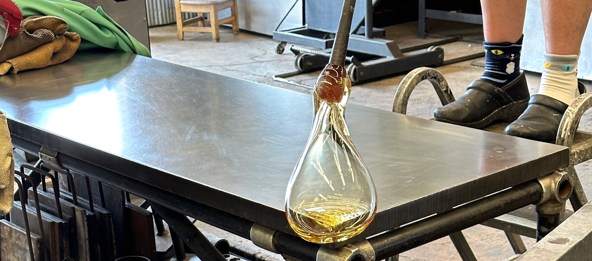 A teardrop-shaped clear glass form hangs from a metal pole over the edge of a metal table. The person holding the pole is partially visible.