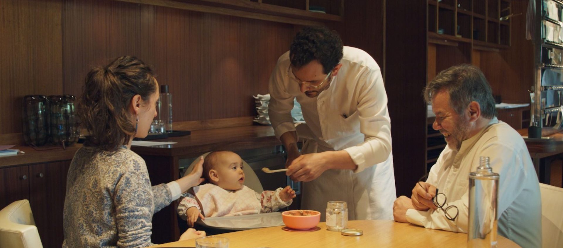 An infant is spoon fed by a chef as the child’s mother and another chef look on.