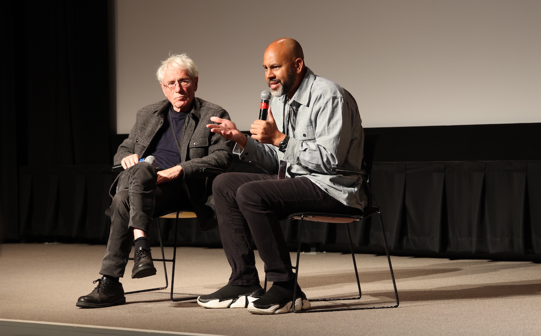 An older man with white hair looks on as a man with brown skin sitting next to him speaks into a microphone.