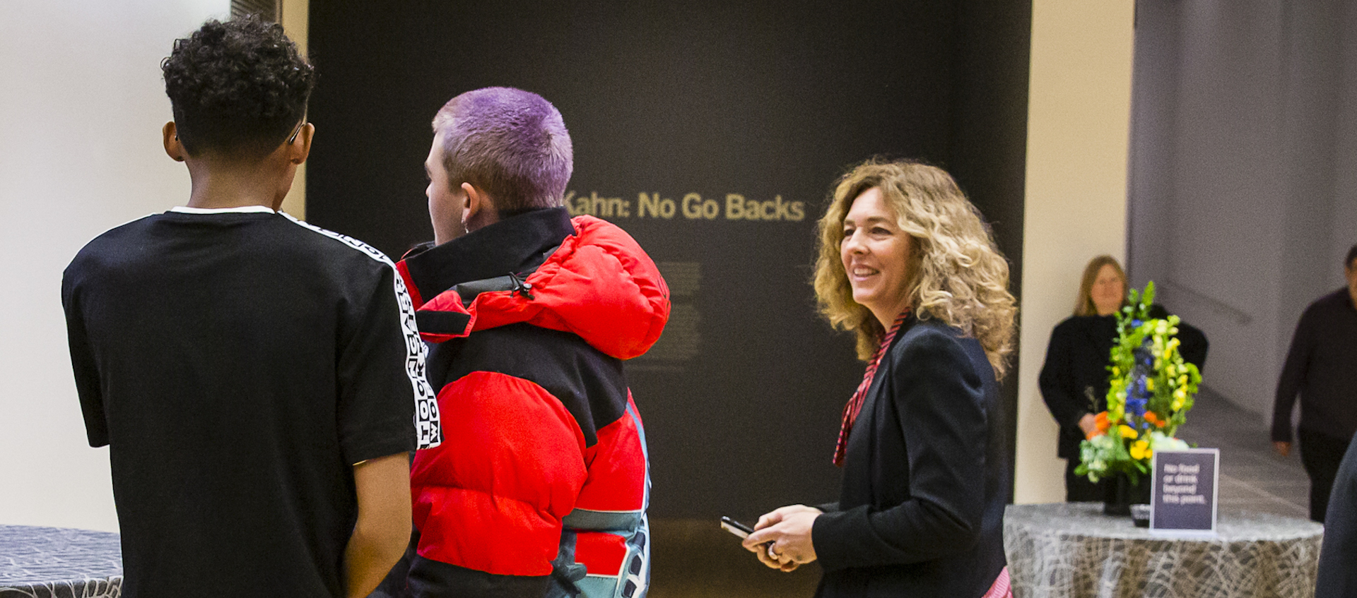 A woman stands at the entrance to a gallery and looks toward two young men with their backs to the camera.