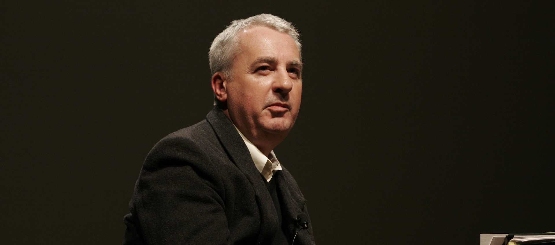 A man with short white hair, wearing a dark suit jacket, looks forward at an unseen audience.