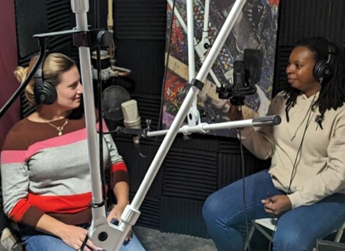 Two women look at each other and smile as they sit together in a podcast recording studio.