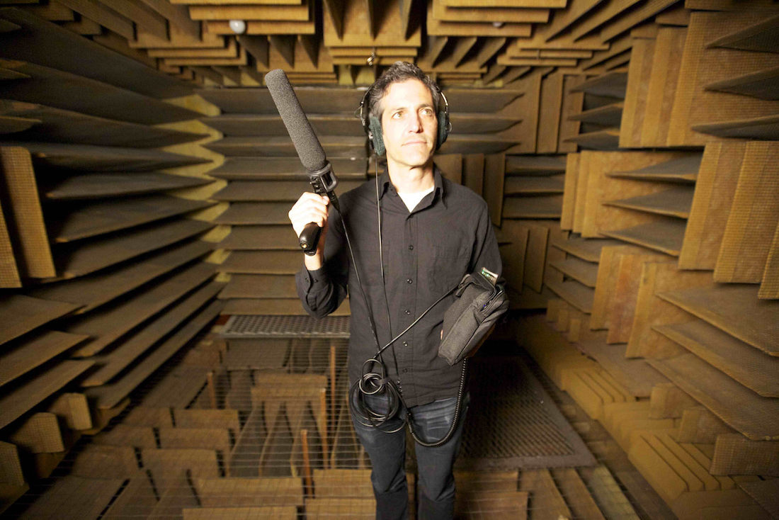 A man wears headphones and holds a boom microphone inside a small room covered in sound-dampening tiles.