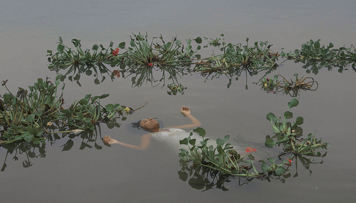 A woman with brown skin wears a white dress and floats in still water with her arms outstretched. Leafy water plants with small flowers surround her.