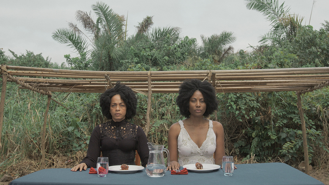 Two Black women sit side by side outside at a table set for a meal amid tropical vegetation. One wears black, the other wears white, both stare ahead.
