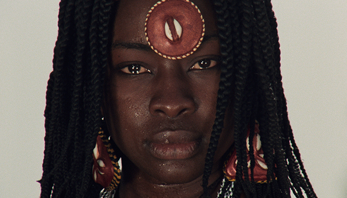 A headshot of a Black woman with long braids wearing a headband with a disc that covers her forehead and matching earrings.