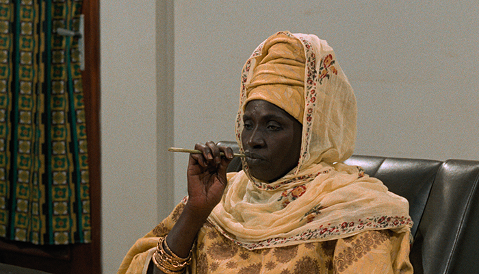 A woman wearing a bright yellow headdress and scarf is sitting in a chair smoking a long pipe. 