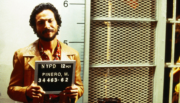 Benjamin Bratt as a bearded Piñero wears a leather jacket, smirks, and holds a placard that says "NYPD 12 PCT, Pinero, M" for his mugshot.