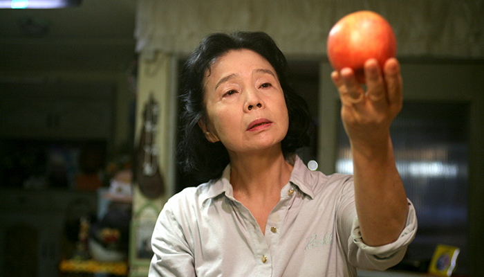 An older Korean woman stands in her kitchen. Facing the camera, she looks closely at an red apple streaked with yellow that she holds up toward the light.