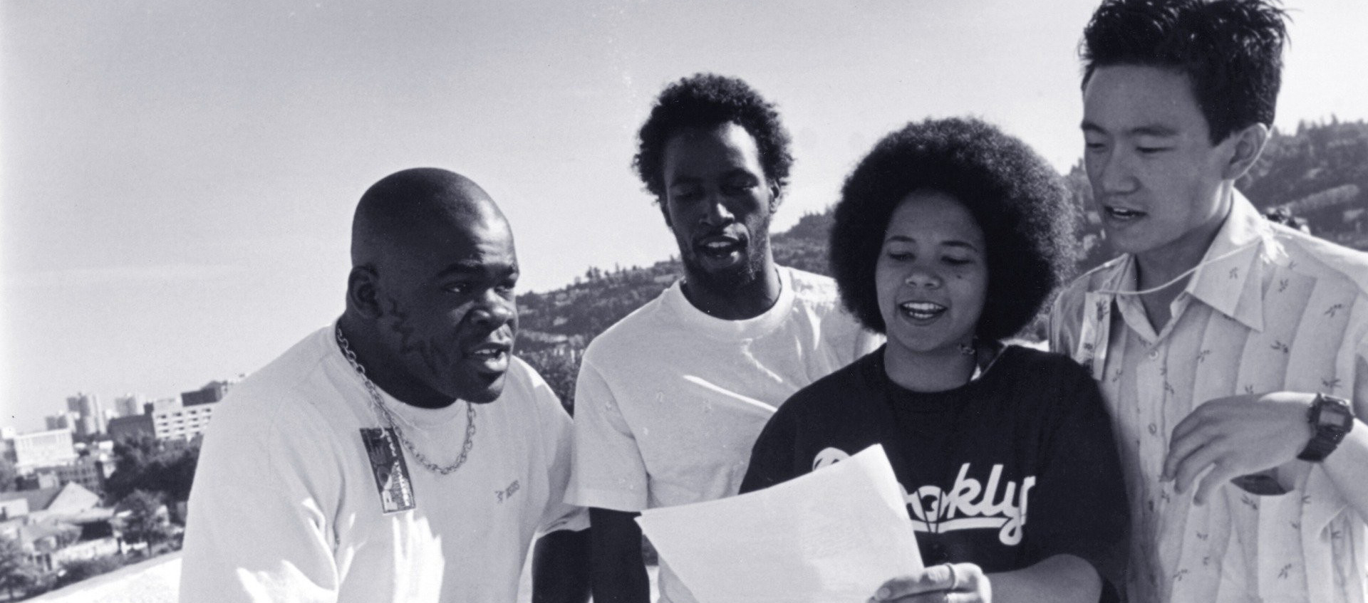 Two young Black men, a young Asian man, and a young Black woman stand together outside and read aloud from a sheet of paper the woman holds in front of her.