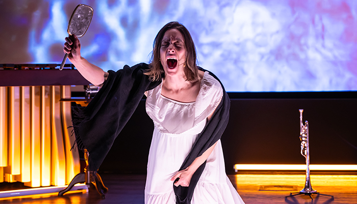 An actor in a white dress and black scarf holds a mirror in the air while screaming.