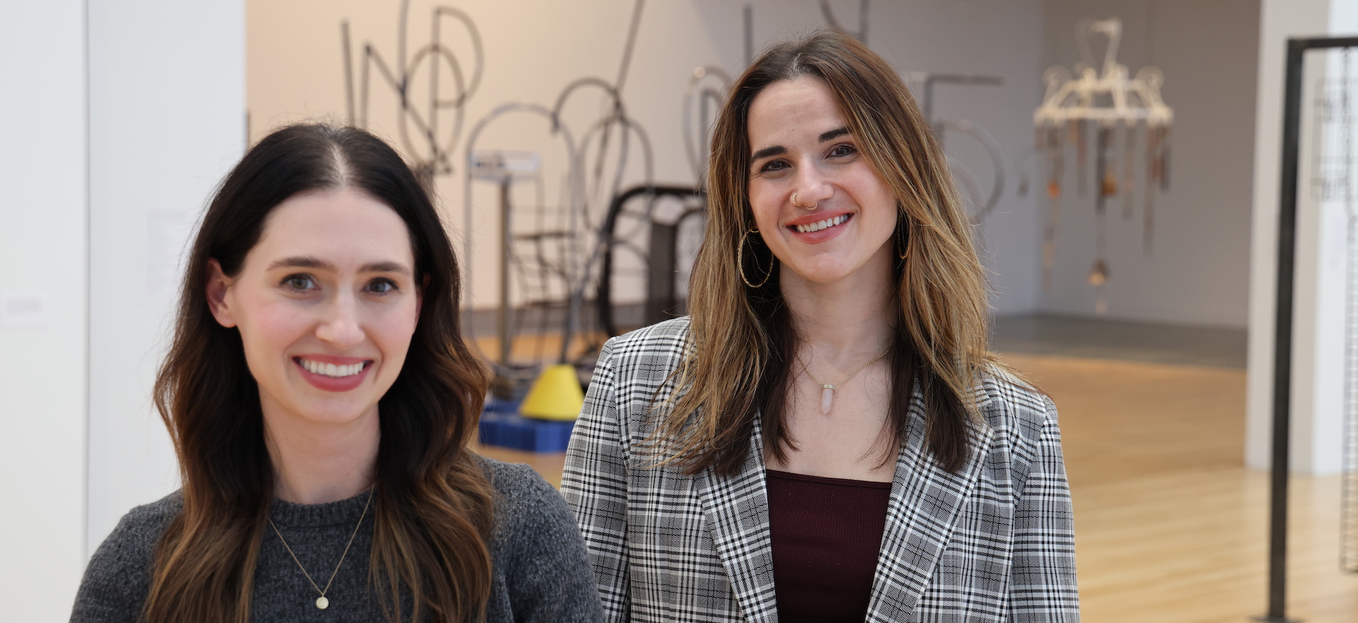 Two women stand together and smile in a gallery with artworks installed behind them.