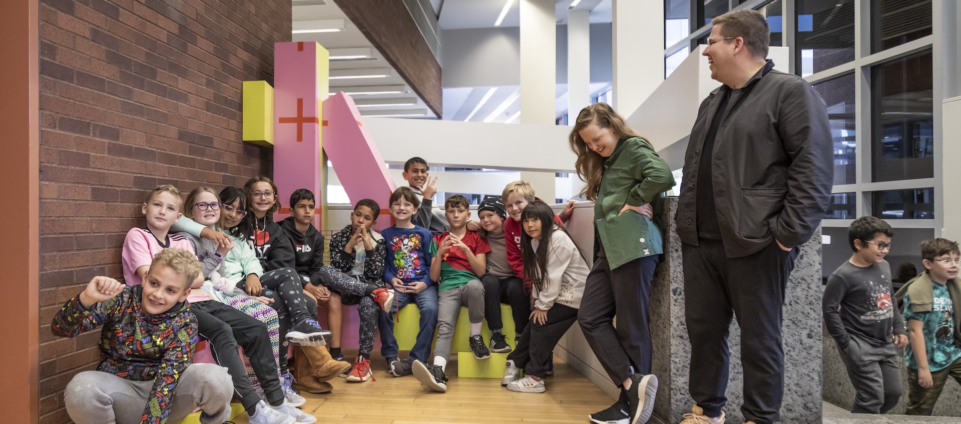 A group of grade school students sit in a contemporary museum lobby on a large, colorful piece of modular furniture.